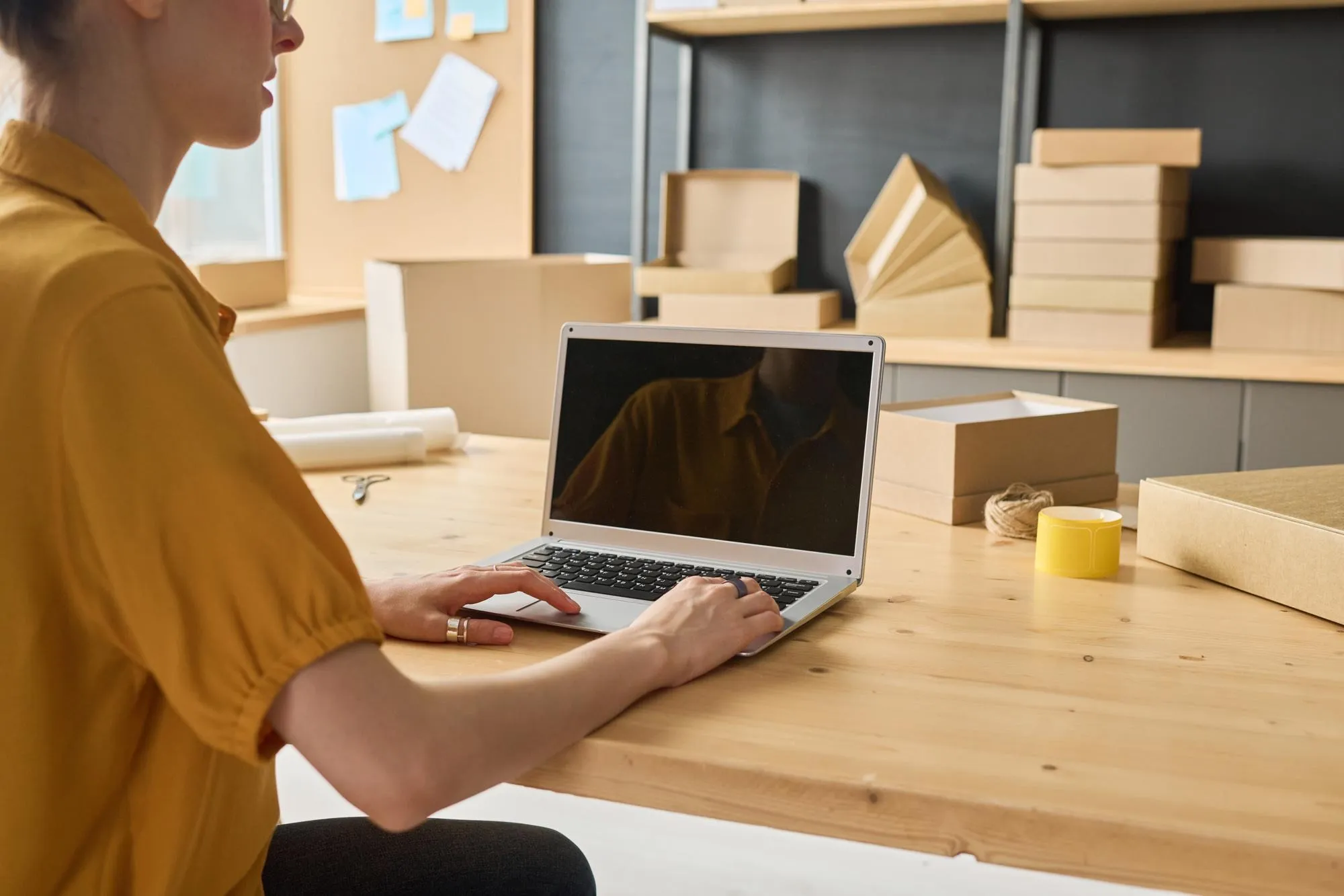 Onlinehändlerin am Packtisch arbeitet am Laptop zwischen Versandkartons